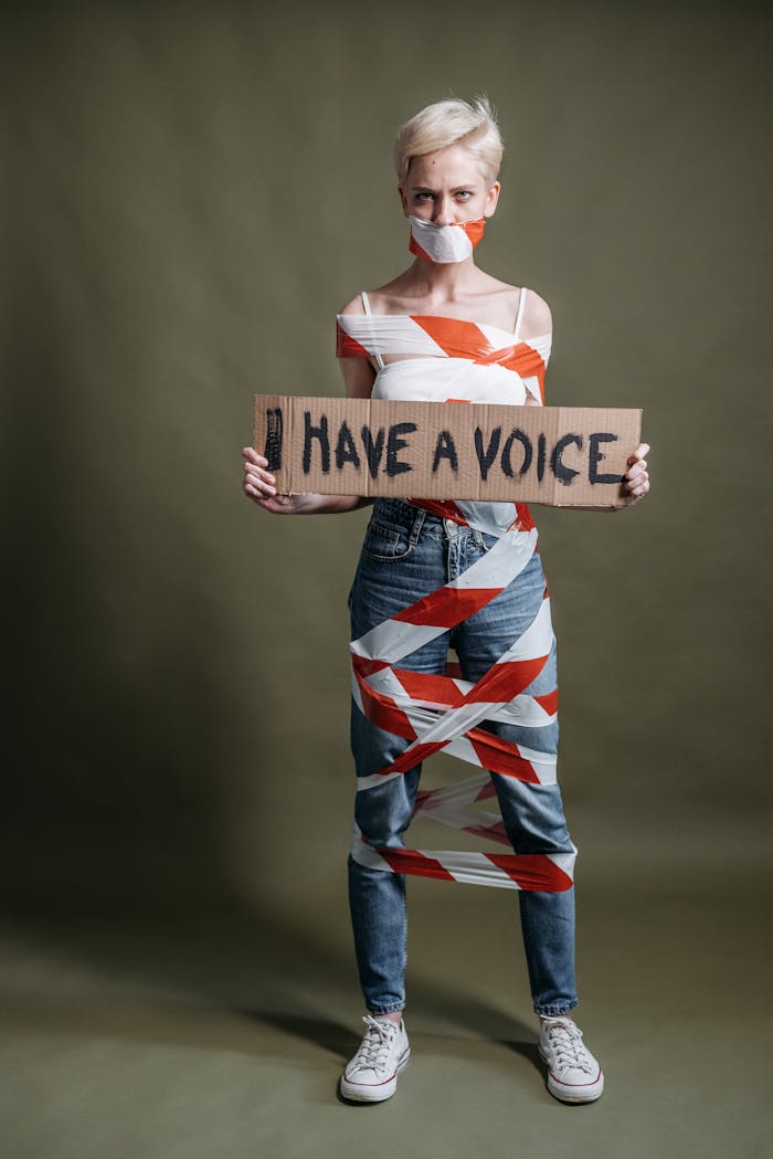 Woman wrapped in tape holding a sign stating 'I Have a Voice' against a studio backdrop.