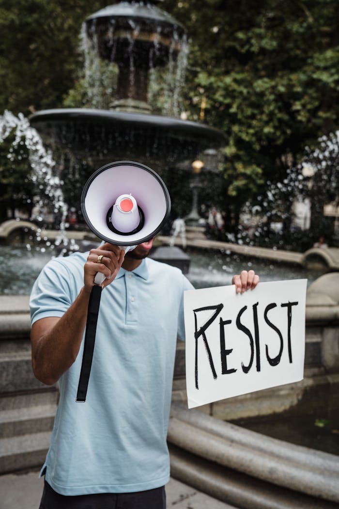 A protester holding a sign reading 'Resist' with a megaphone near a fountain outdoors.