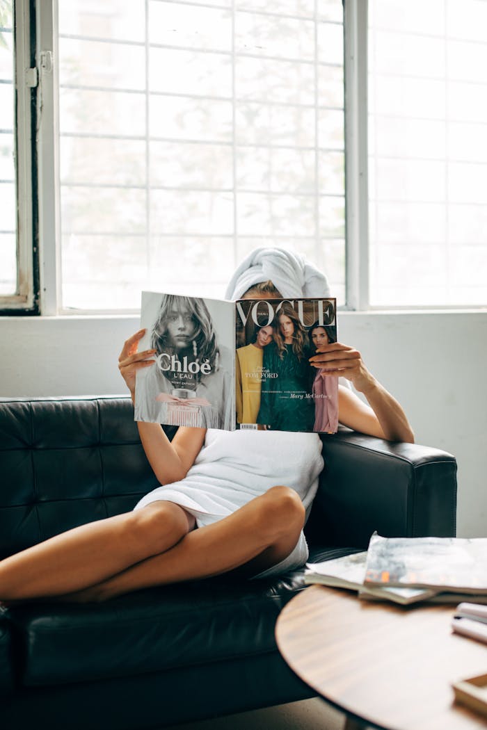 who-we-are A woman with a towel wrapped on her head reads a Vogue magazine while lounging on a couch.